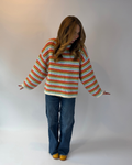 A woman wearing a fuzzy, oversized striped sweater with blue jeans and yellow shoes, standing against a plain light gray studio background.