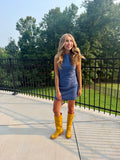 A woman stands outdoors on a concrete patio wearing a sleeveless slate blue mini dress and bright yellow knee-high boots, with a black metal railing and green trees in the background.