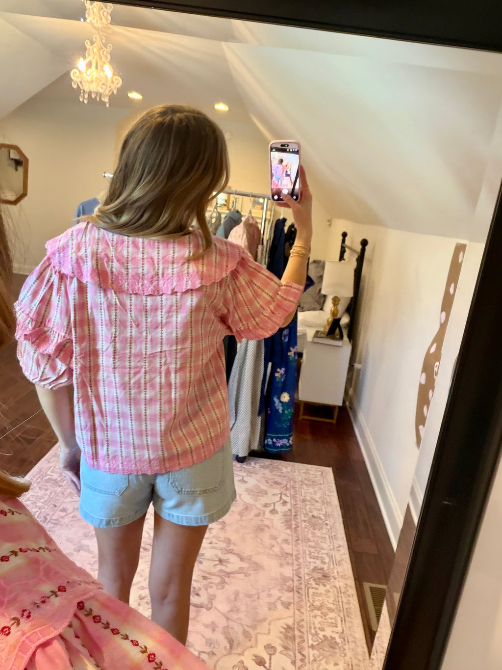 Woman taking a mirror selfie wearing a pink plaid shirt and denim shorts in a room with a chandelier and clothing rack.