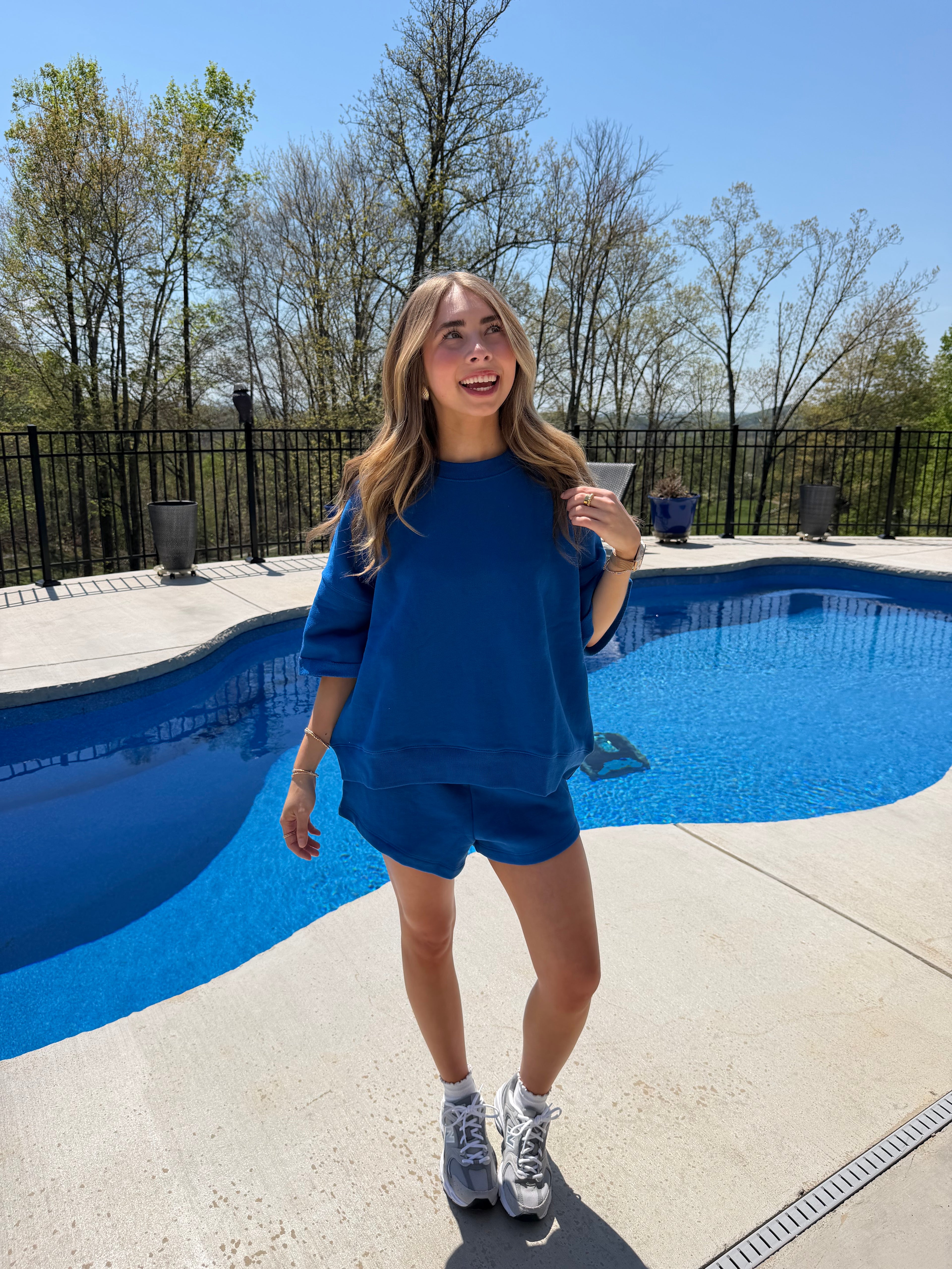 Person in a blue outfit standing near a pool with trees and a fence in the background