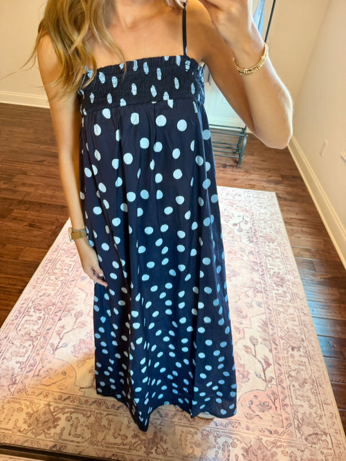 A woman wearing a navy blue maxi dress with a light blue polka dot pattern, taking a mirror selfie in a room with hardwood floors and a patterned rug.