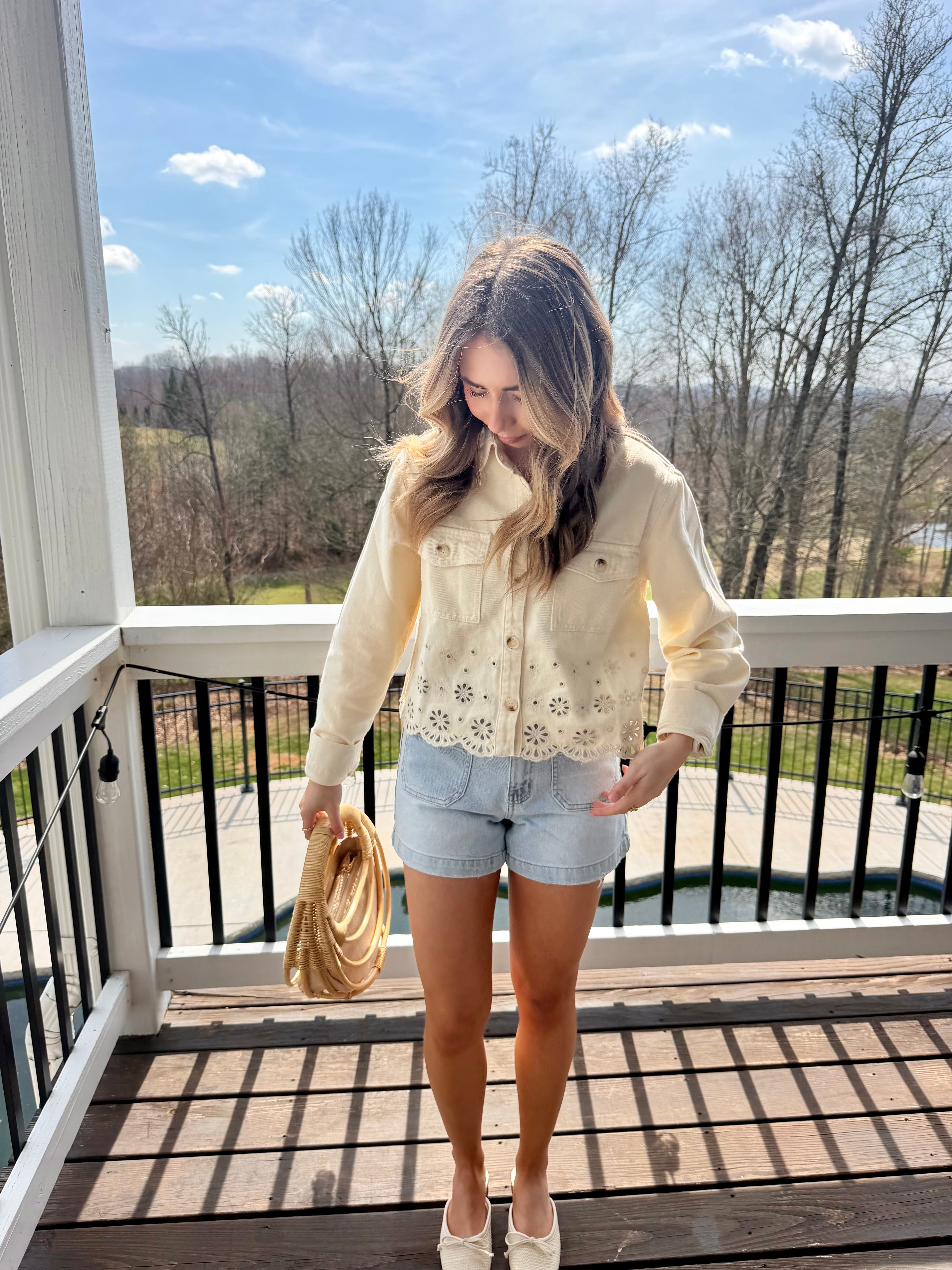 Woman standing on a balcony wearing a light-colored lace top and denim shorts, holding a beige handbag.