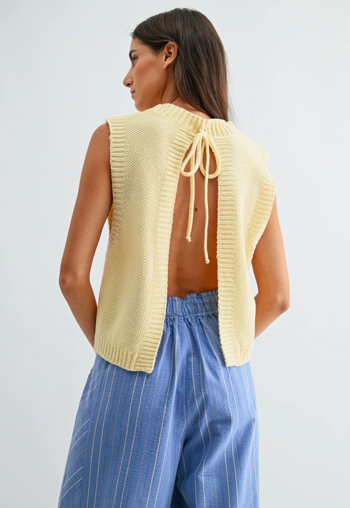 Woman wearing a yellow knit vest with open back and blue striped pants on a light gray background