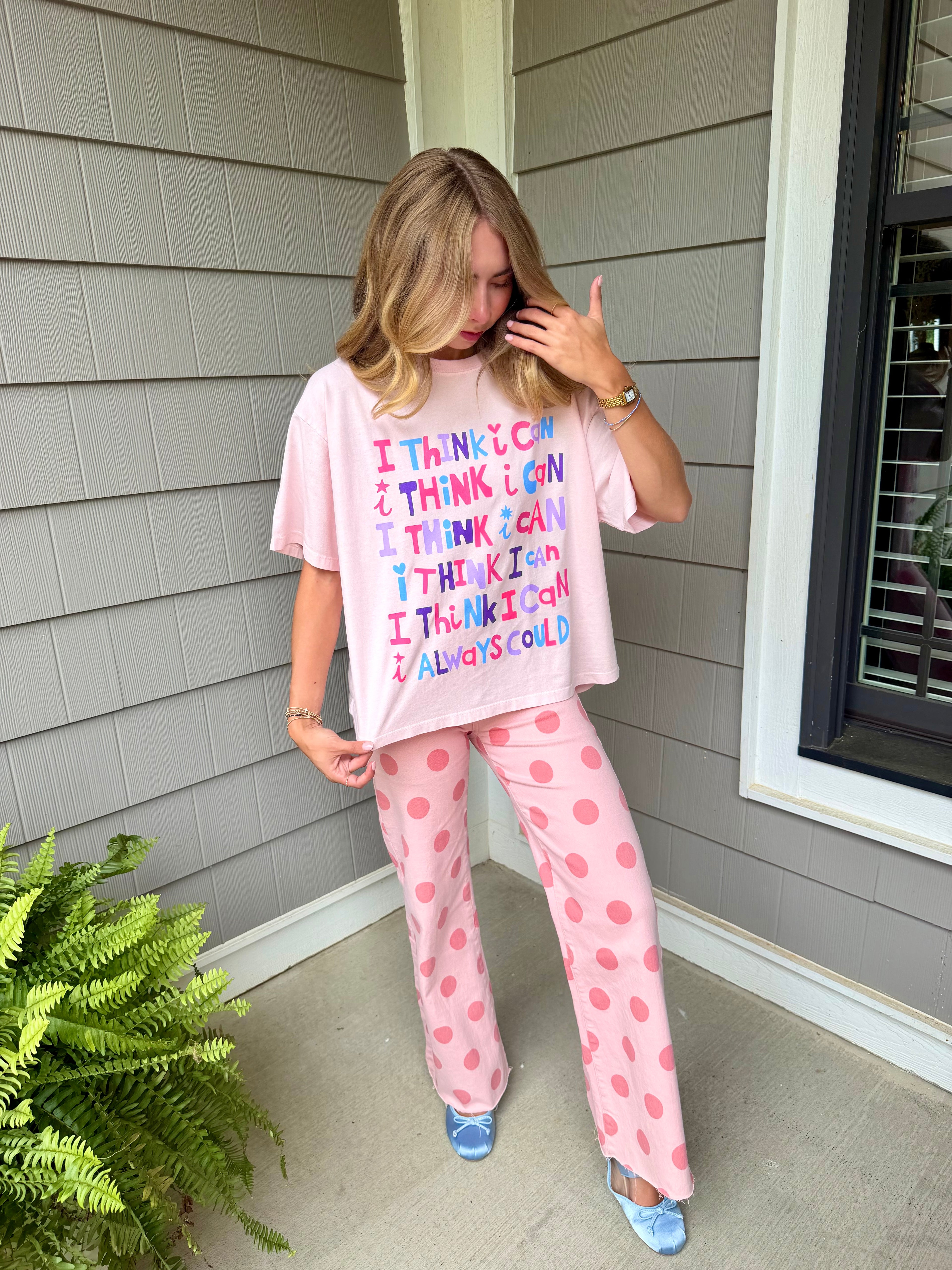 A woman stands on a porch wearing a short-sleeve T-shirt with colorful text reading 'I THINK I CAN I THINK I CAN I THINK I CAN I THINK I CAN I ALWAYS COLD' and matching pink pants with darker pink polka dots.