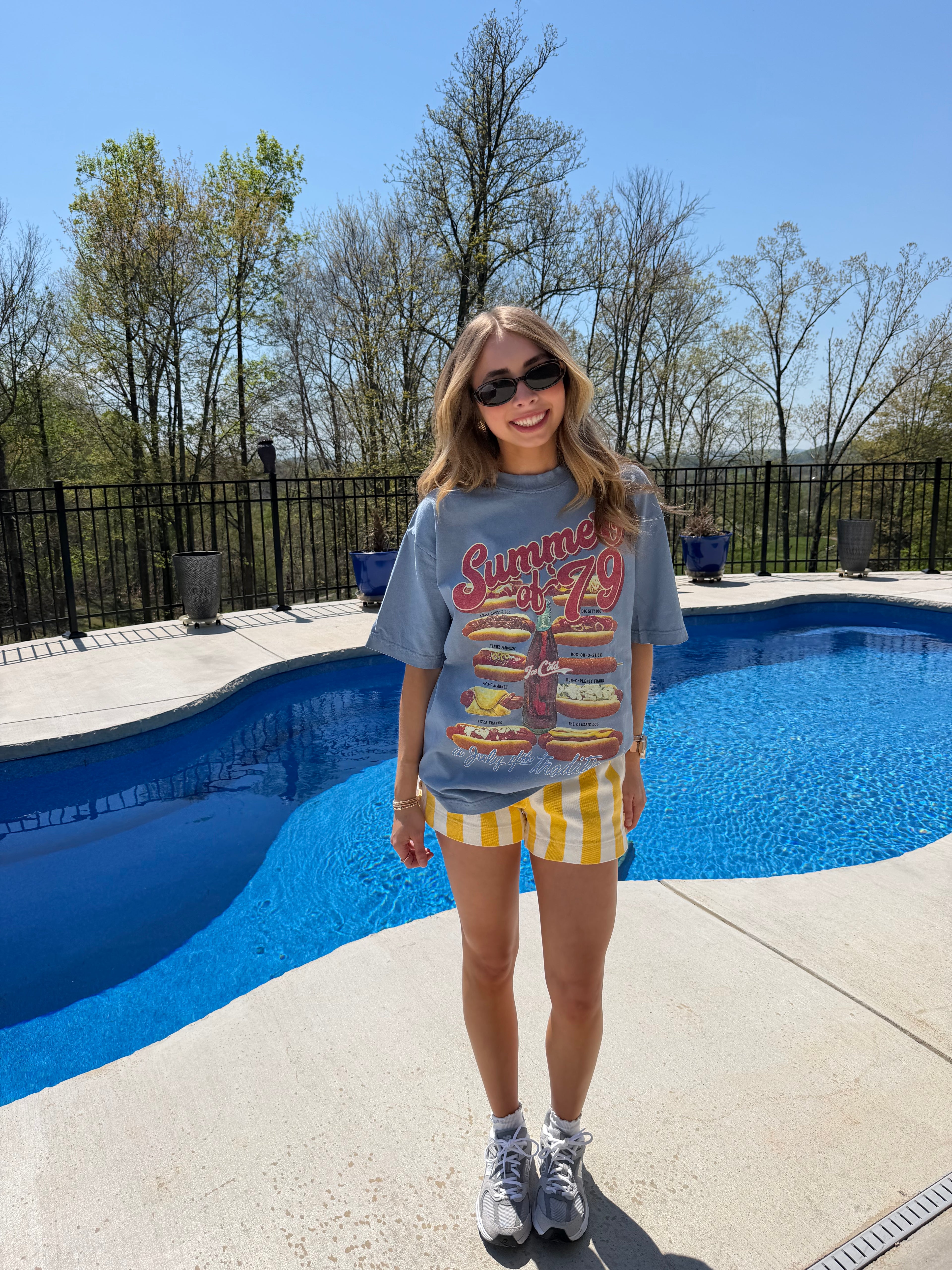Person standing by a pool wearing sunglasses and a graphic t-shirt.