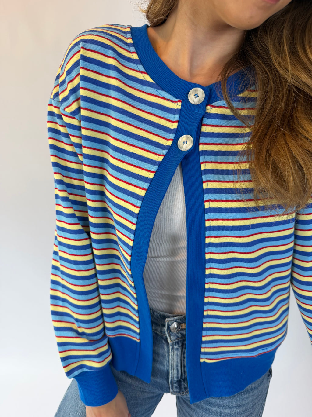 A close-up of a person wearing a blue cardigan with yellow and red horizontal stripes, featuring a solid blue ribbed collar, cuffs, and hem, styled with a white top and blue jeans.