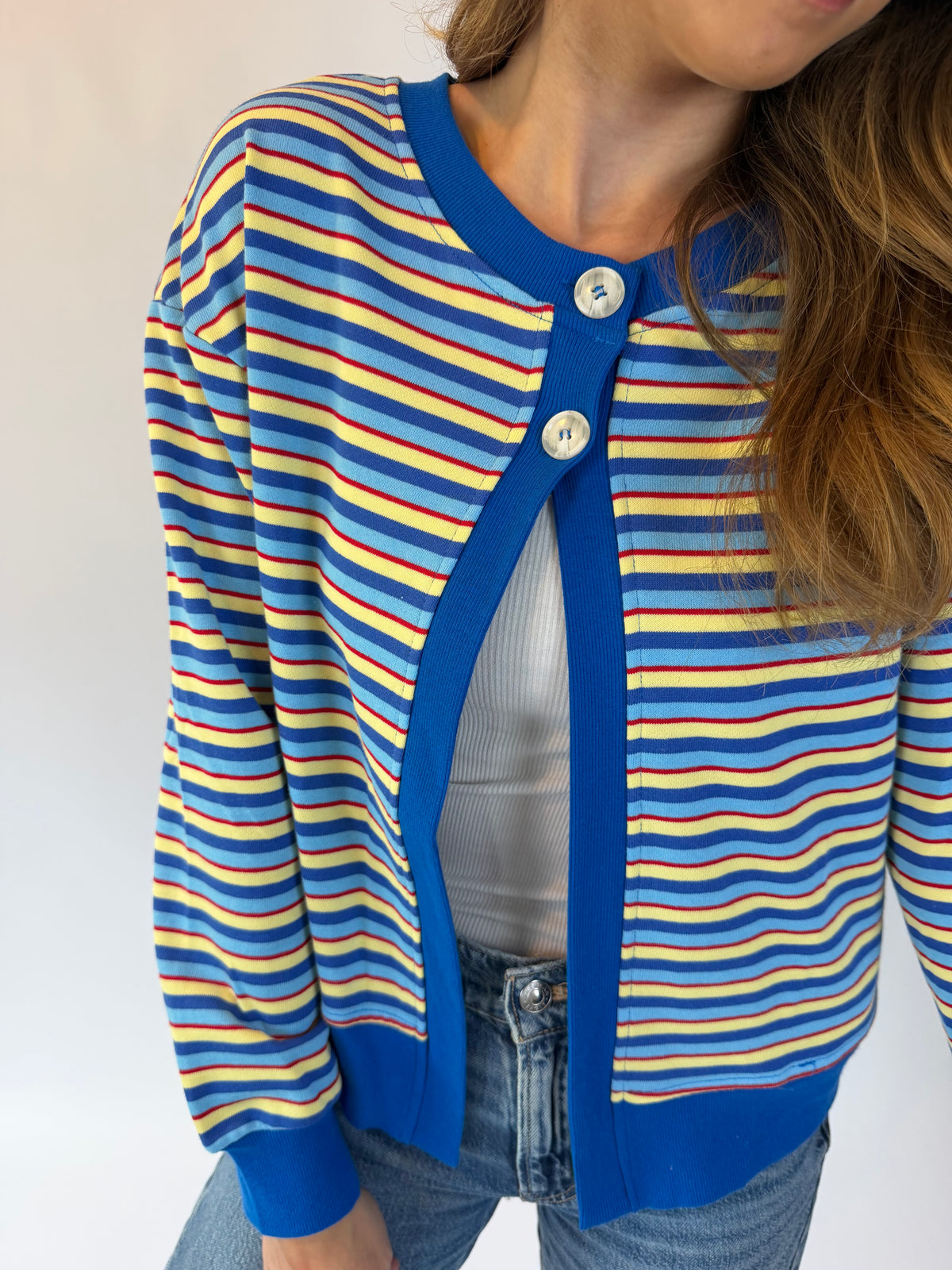 A close-up of a person wearing a blue cardigan with yellow and red horizontal stripes, featuring a solid blue ribbed collar, cuffs, and hem, styled with a white top and blue jeans.