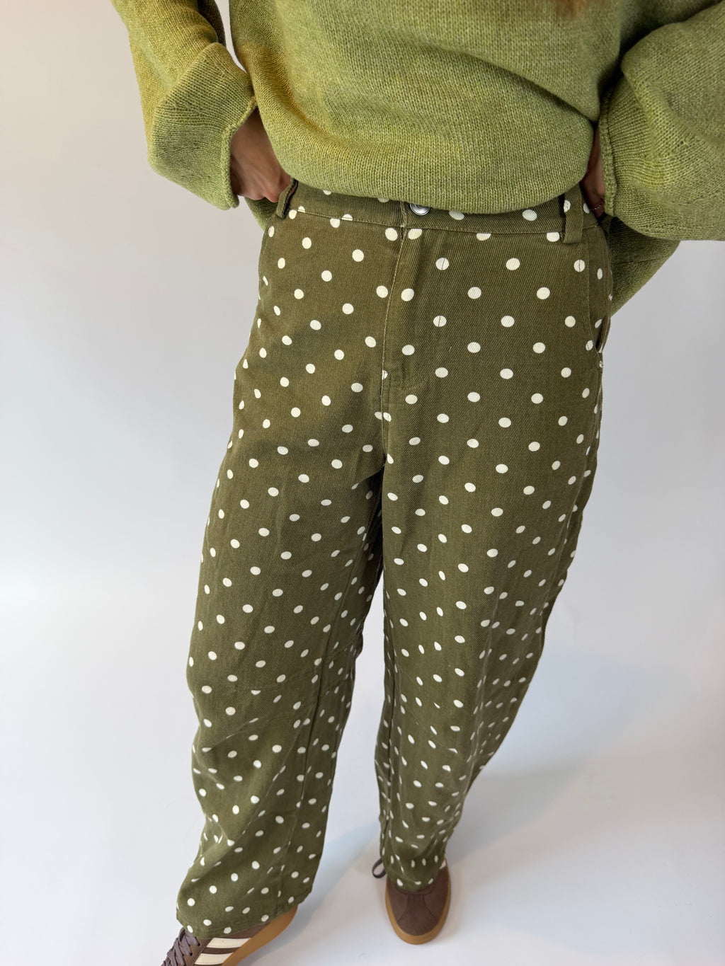 A person wearing olive green pants with a white polka dot pattern, styled with a matching olive green knit sweater and brown shoes, photographed against a plain white studio background.