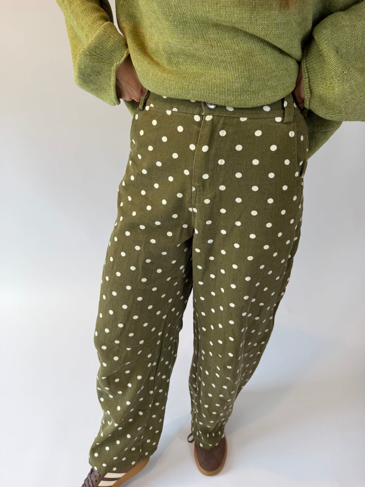 A person wearing olive green pants with a white polka dot pattern, styled with a matching olive green knit sweater and brown shoes, photographed against a plain white studio background.