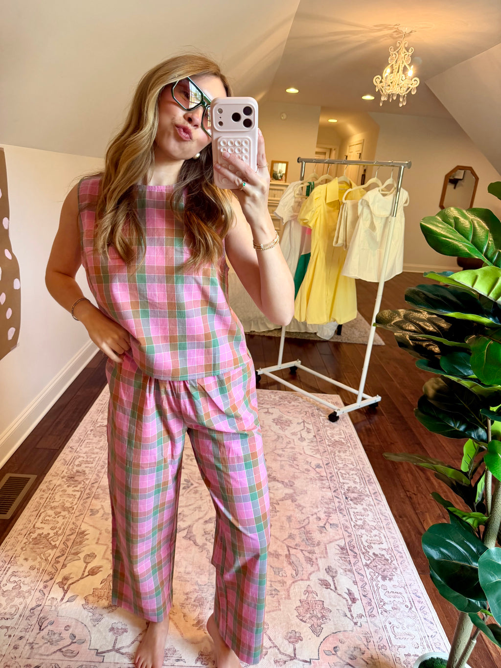 Woman taking a mirror selfie wearing a pink plaid outfit in a room with a plant and clothing rack.