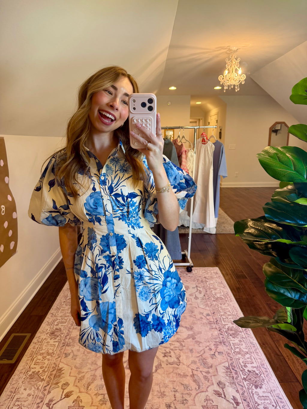 Woman taking a mirror selfie wearing a blue floral dress in a room with plants and a chandelier.