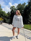 A woman wearing an oversized light gray sweatshirt with 'PRAY' printed in black-and-white patterned letters, standing outdoors on a concrete patio with trees and a black railing behind her.