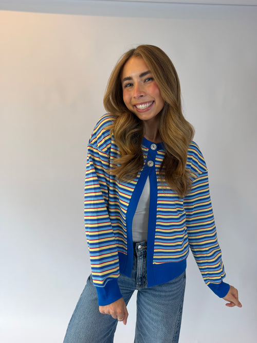 A woman modeling a blue, yellow, and white striped knit cardigan with a solid blue front placket, worn over a gray top and blue jeans against a plain white studio background.