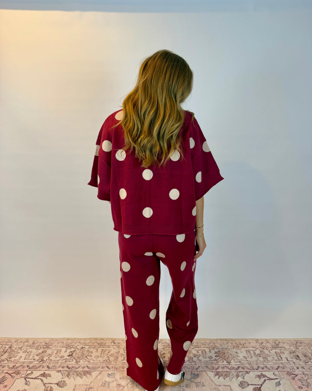 A woman viewed from behind wearing a matching burgundy two-piece pajama set with large white polka dots, consisting of a short-sleeve top and wide-leg pants, standing against a plain white wall.