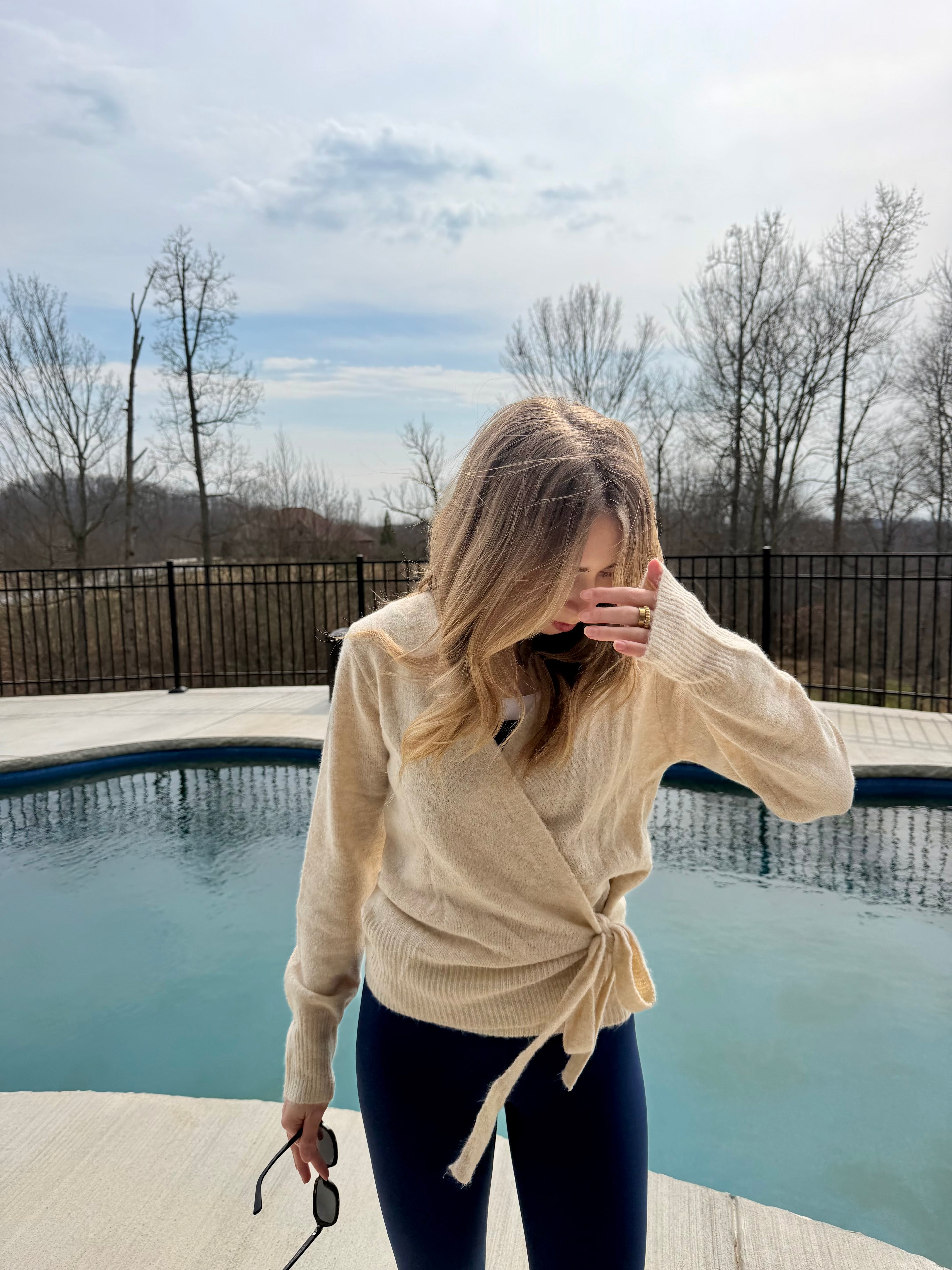 Woman in a beige cardigan standing by a pool with trees and a cloudy sky in the background