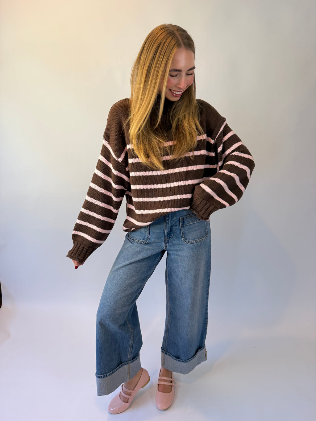 A woman wearing a brown and pink striped knit sweater, blue jeans, and pink ballet flats, photographed against a plain light background.