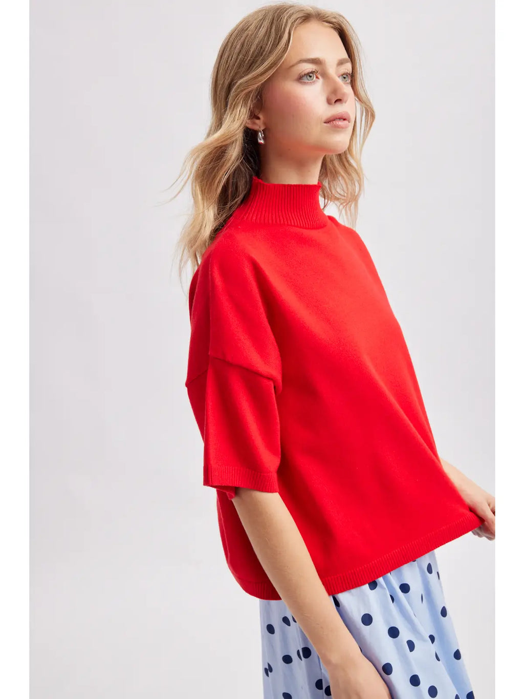 A woman wearing a bright red knit mock-neck top with short sleeves and light blue polka-dot pants, photographed against a plain light gray studio background.