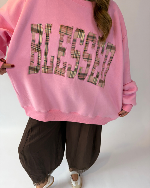 A person wearing an oversized pink sweatshirt with the word 'BLESSED' in large plaid-patterned letters, paired with dark brown harem-style pants and white shoes, against a plain light background.