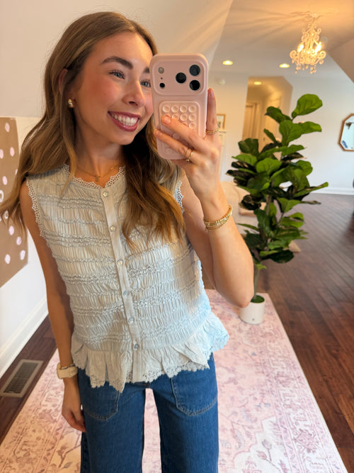 Woman taking a selfie in a room with a plant and decorative items. Wearing a baby blue tank top.
