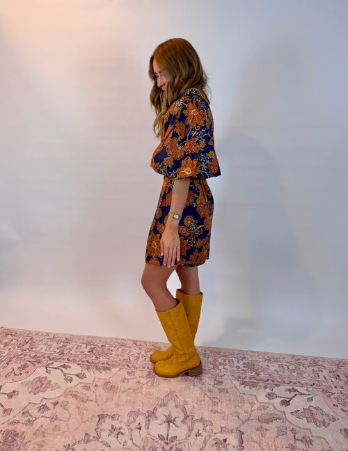 A woman stands in profile wearing a short floral dress with wide flutter sleeves and mustard yellow knee-high boots, standing on a patterned rug against a plain light wall.