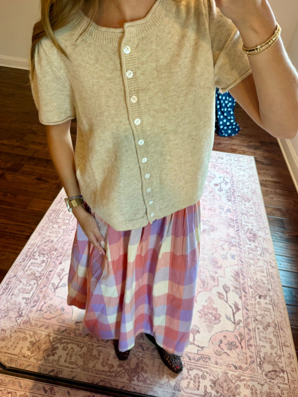 A person wearing a short-sleeve beige knit cardigan with white buttons and a pink, purple, and blue plaid maxi skirt, standing on a patterned rug in a home setting.