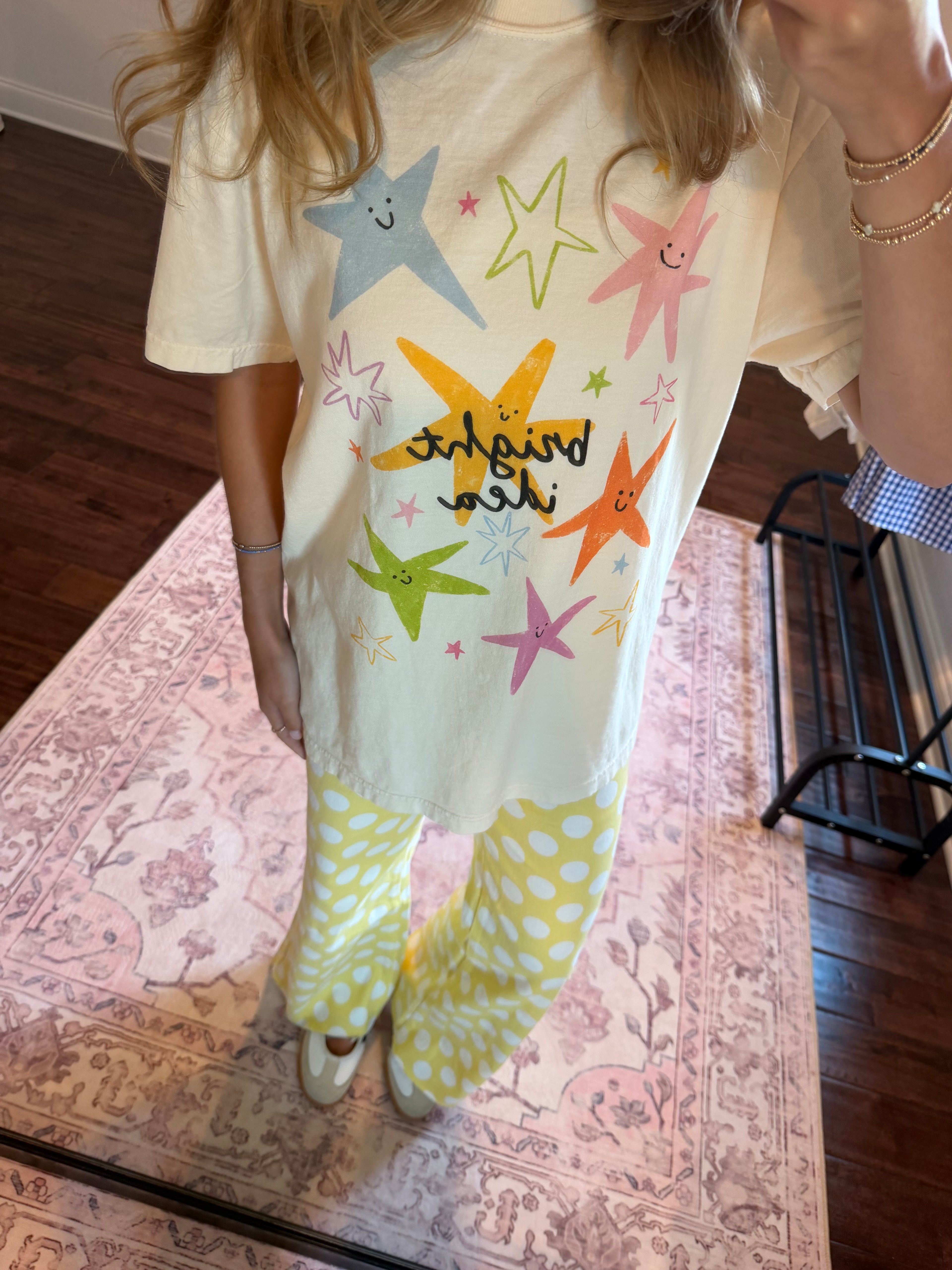 A person wearing a cream-colored graphic t-shirt with colorful star prints and the text 'shine and shine' paired with yellow-and-white polka dot pants, standing on a patterned rug in a home setting.