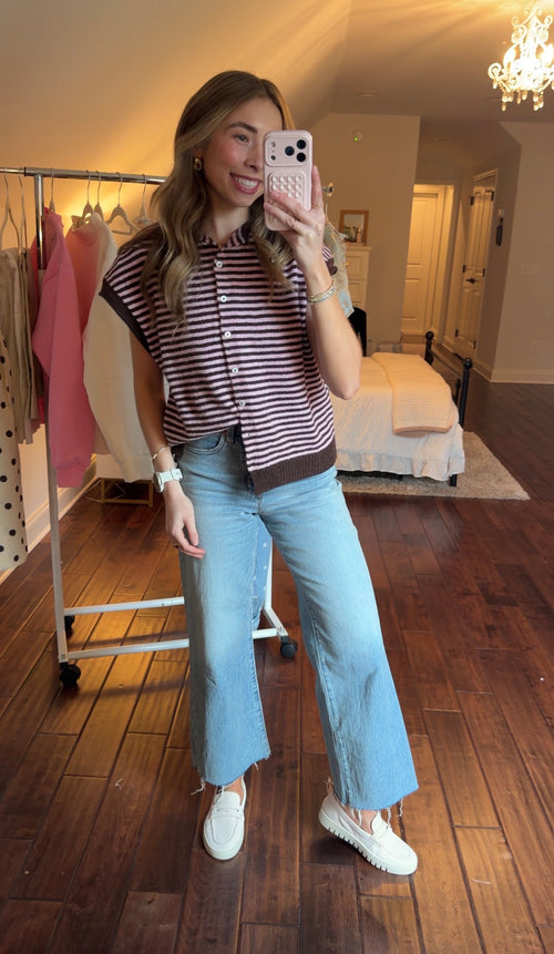 Person taking a mirror selfie wearing a pink and brown striped sweater vest and jeans in a bedroom.