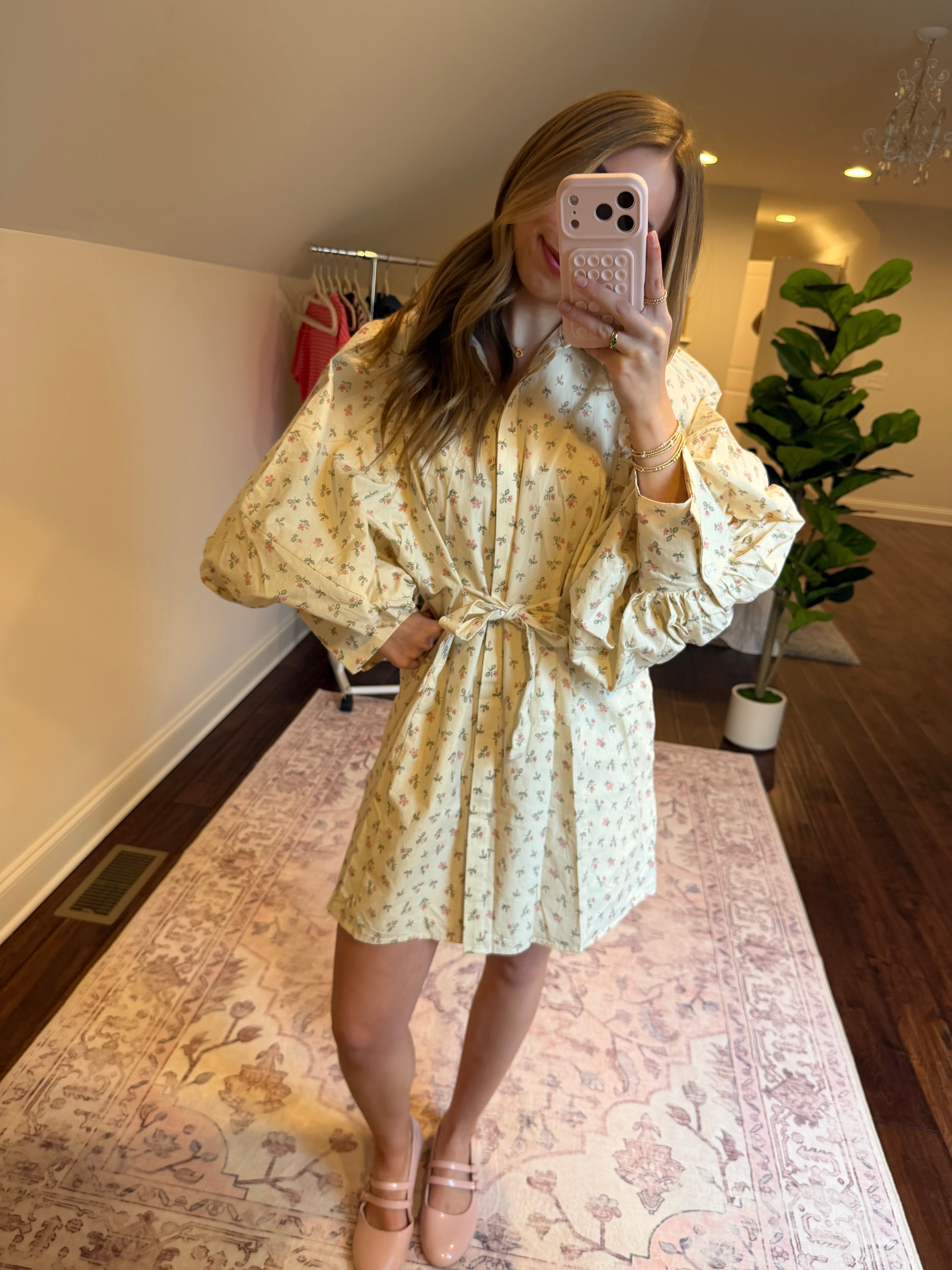 Woman in a floral dress standing on a patterned rug in a room with a plant and furniture.