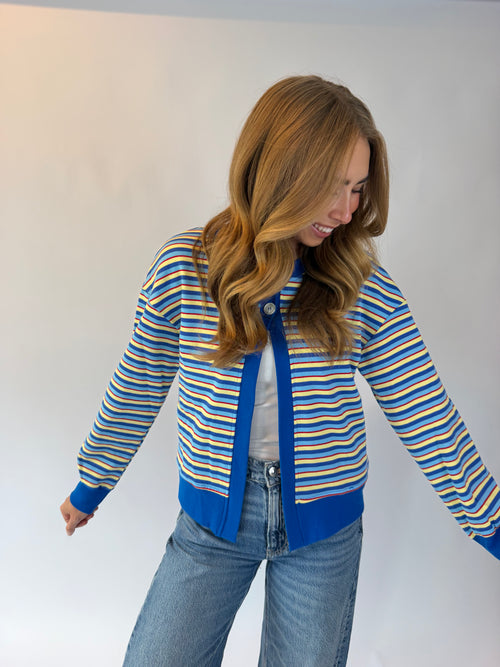 A woman wearing a blue, yellow, and white striped cardigan with solid blue trim, paired with a white t-shirt and light blue jeans, posing against a plain white studio background.