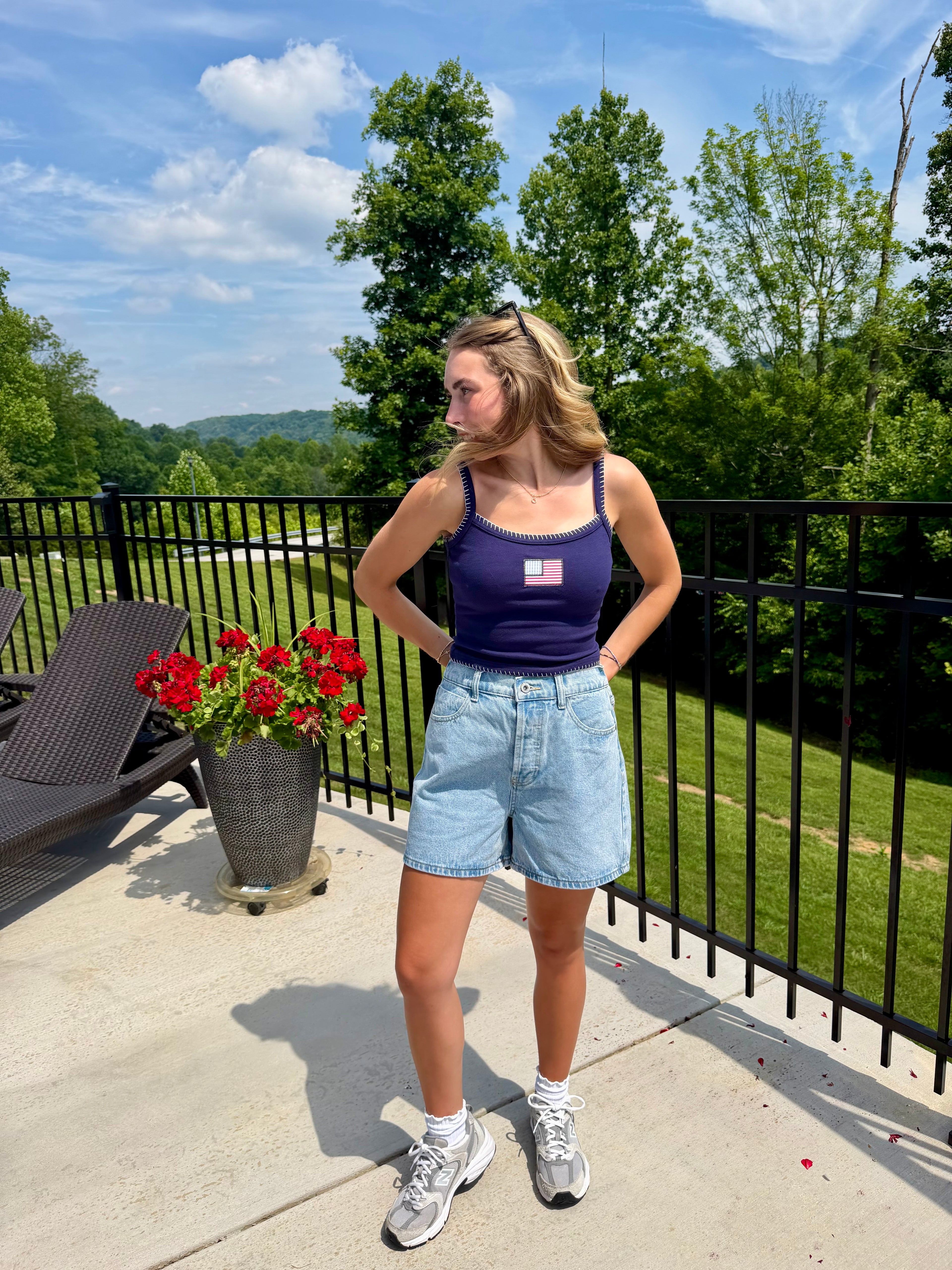 Woman standing on a balcony with a scenic view, wearing a navy tank top and denim shorts.