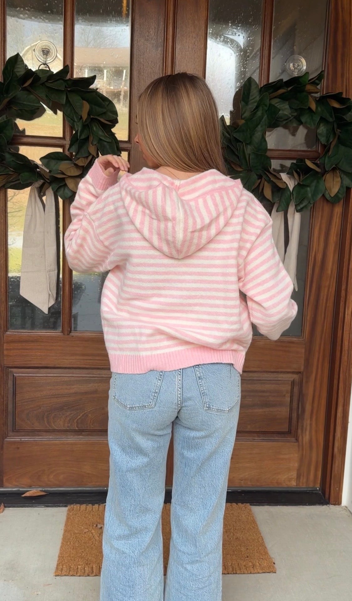 Person wearing a pink striped hoodie and light blue jeans standing in front of a wooden door.