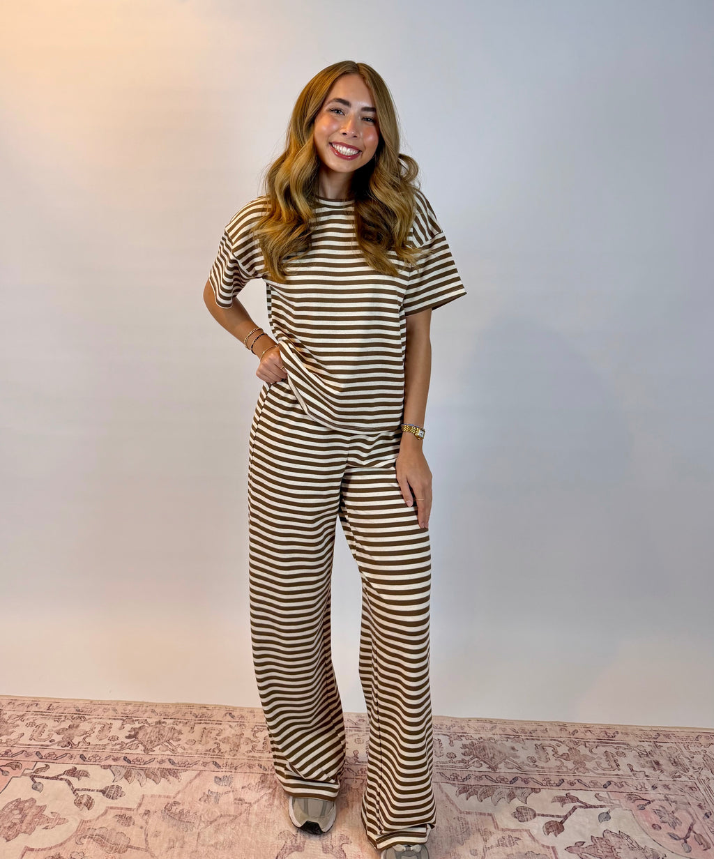 A woman modeling a matching brown and cream striped short-sleeve top and wide-leg pants set, standing on a patterned rug against a plain light gray wall.