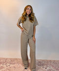 A woman modeling a matching brown and cream striped short-sleeve top and wide-leg pants set, standing on a patterned rug against a plain light gray wall.