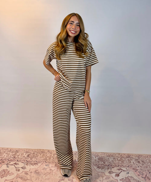 A woman modeling a matching brown and cream striped short-sleeve top and wide-leg pants set, standing on a patterned rug against a plain light gray wall.