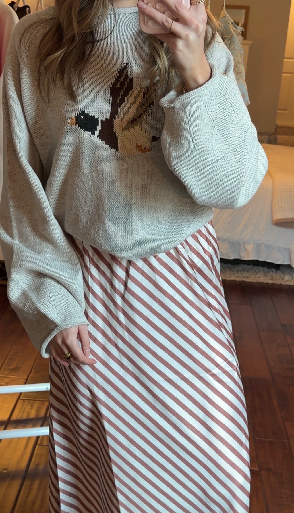 Woman taking a mirror selfie wearing a light-colored sweater with a design (duck design) and a striped skirt in a bedroom.