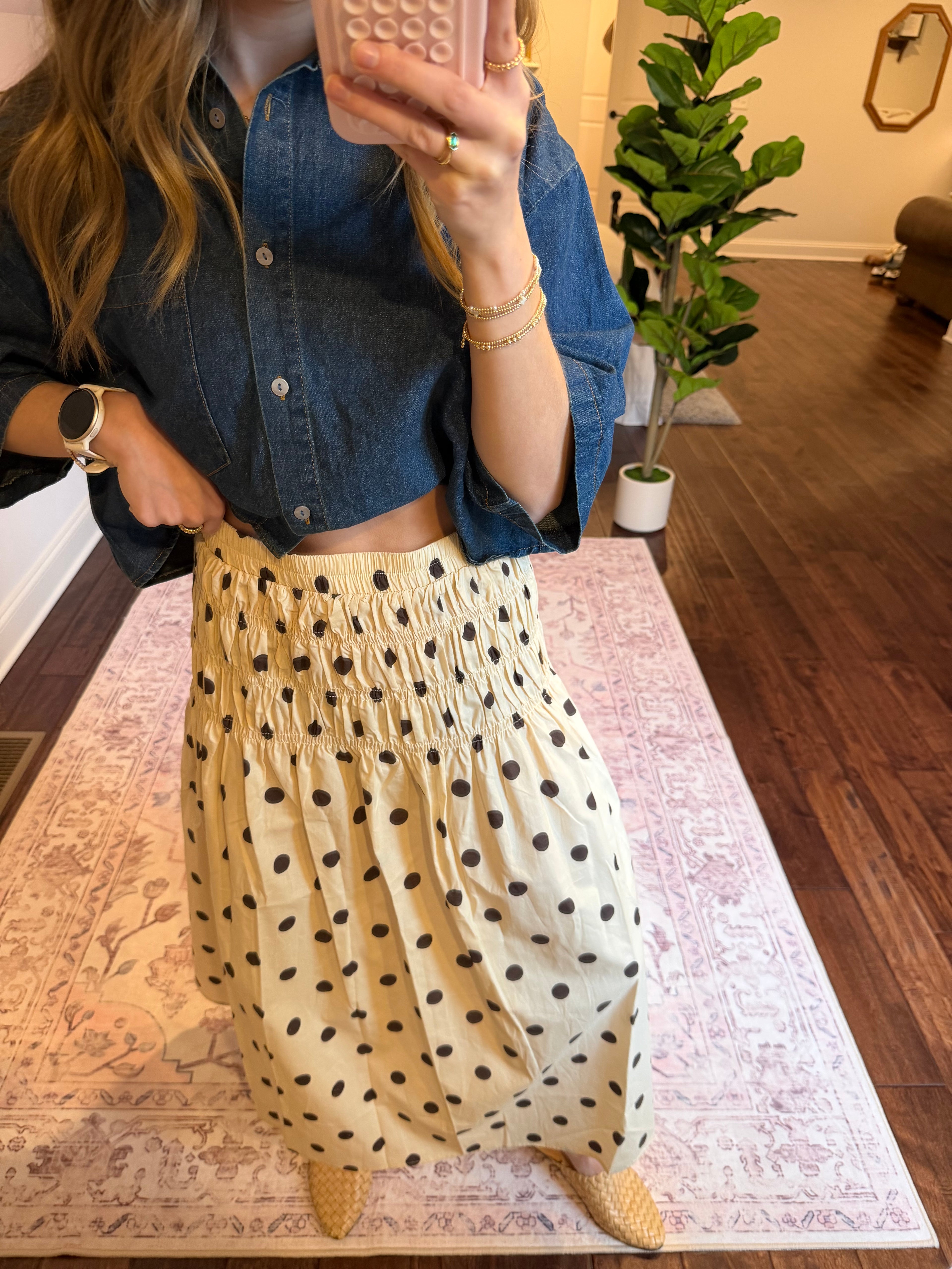 Person with a polka dot skirt and denim top in a room with a plant and wooden floor.