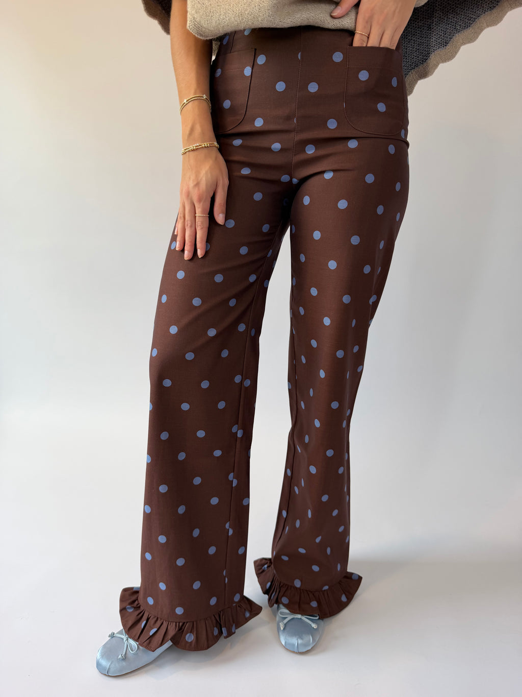 A person wearing high-waisted brown wide-leg pants with a light blue polka dot pattern and ruffled hems, paired with light blue flats, against a plain light background.