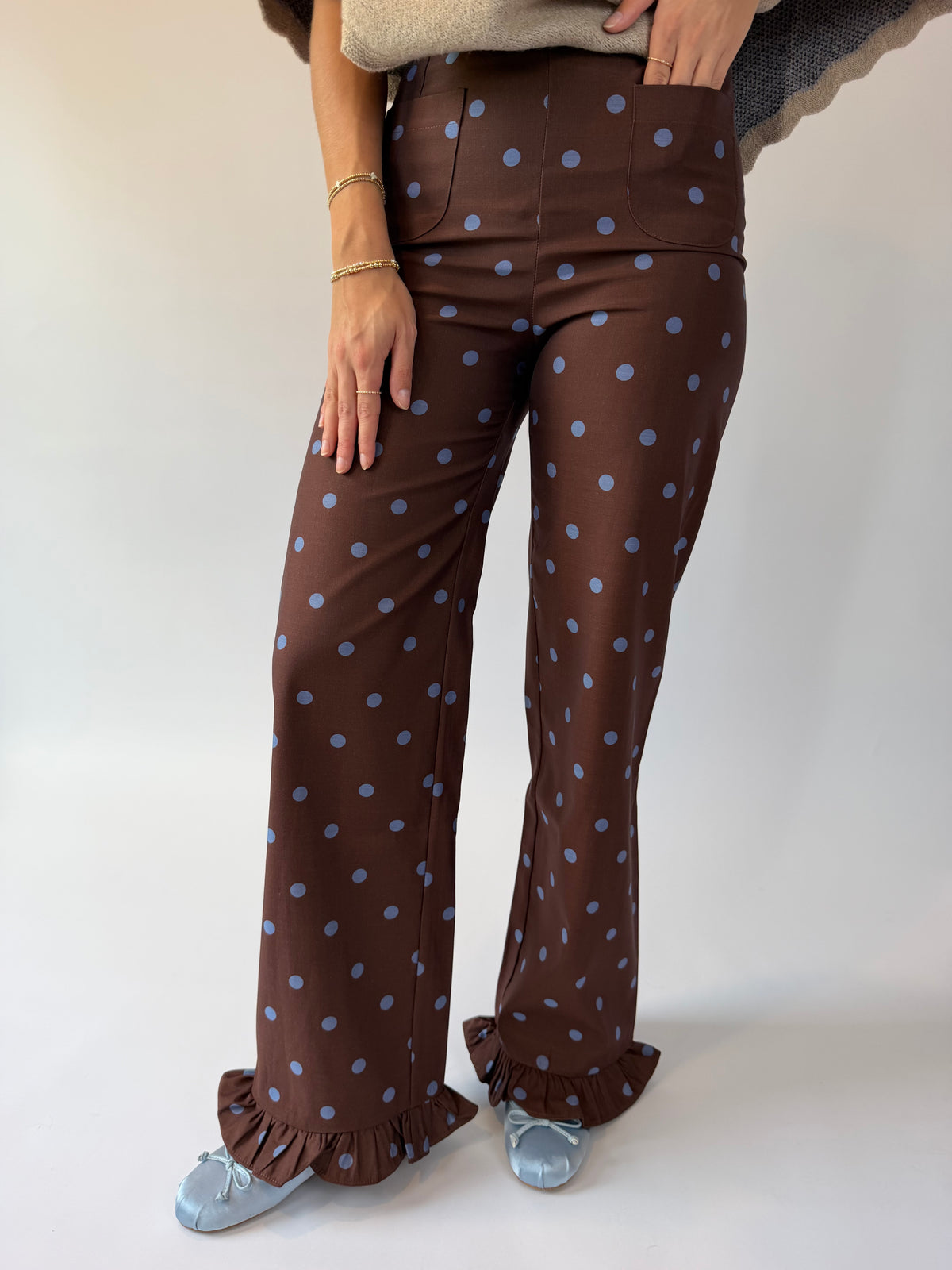 A person wearing high-waisted brown wide-leg pants with a light blue polka dot pattern and ruffled hems, paired with light blue flats, against a plain light background.