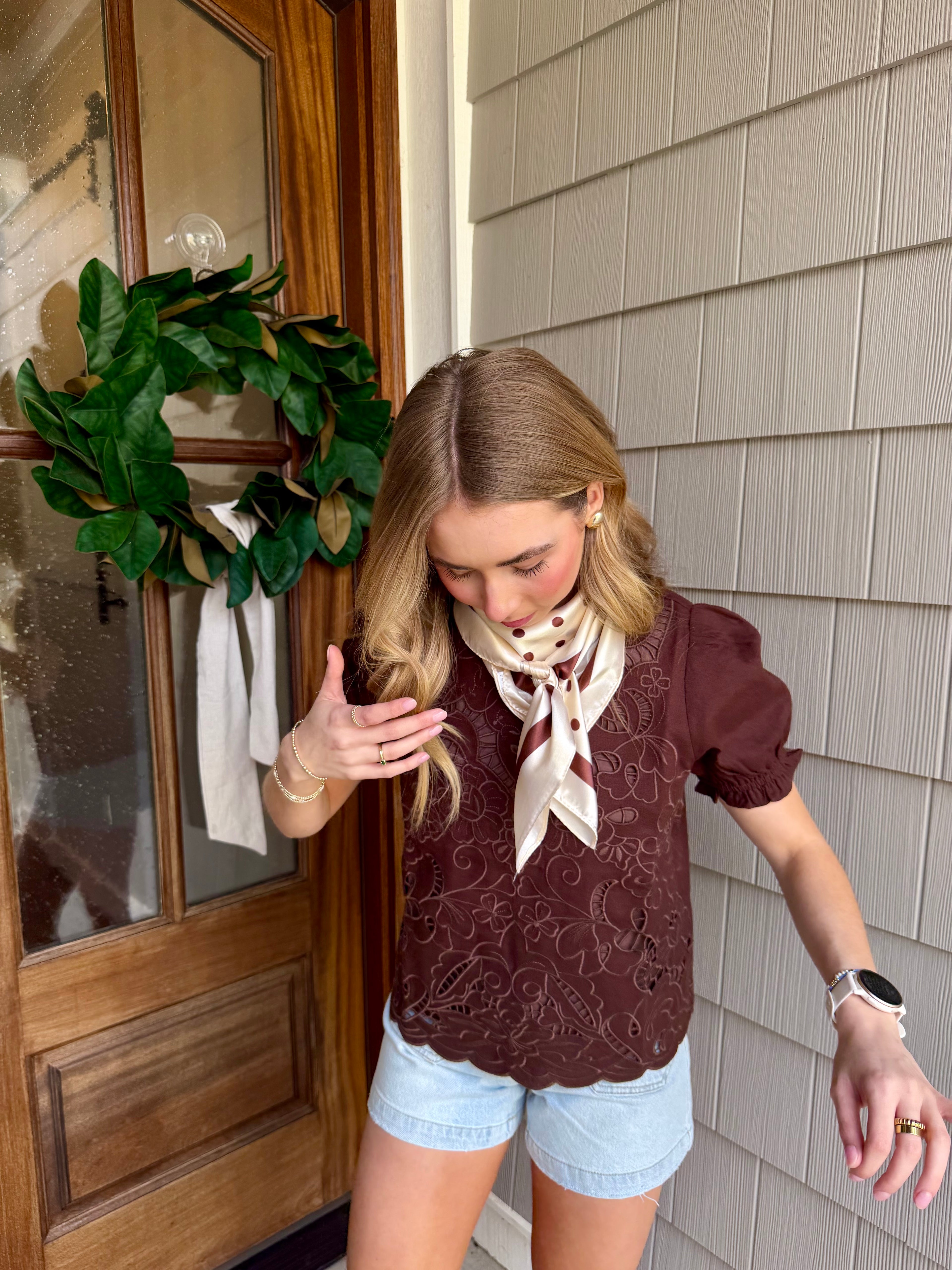 Woman standing outside a house with a wreath on the door, wearing a brown top and light blue shorts.