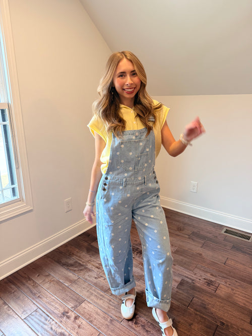 Woman wearing a yellow top and light blue polka dot overalls in a room with wooden flooring.