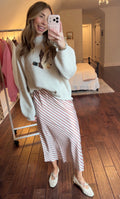 Woman taking a mirror selfie wearing a light-colored sweater with a design (duck design) and a striped skirt in a bedroom.