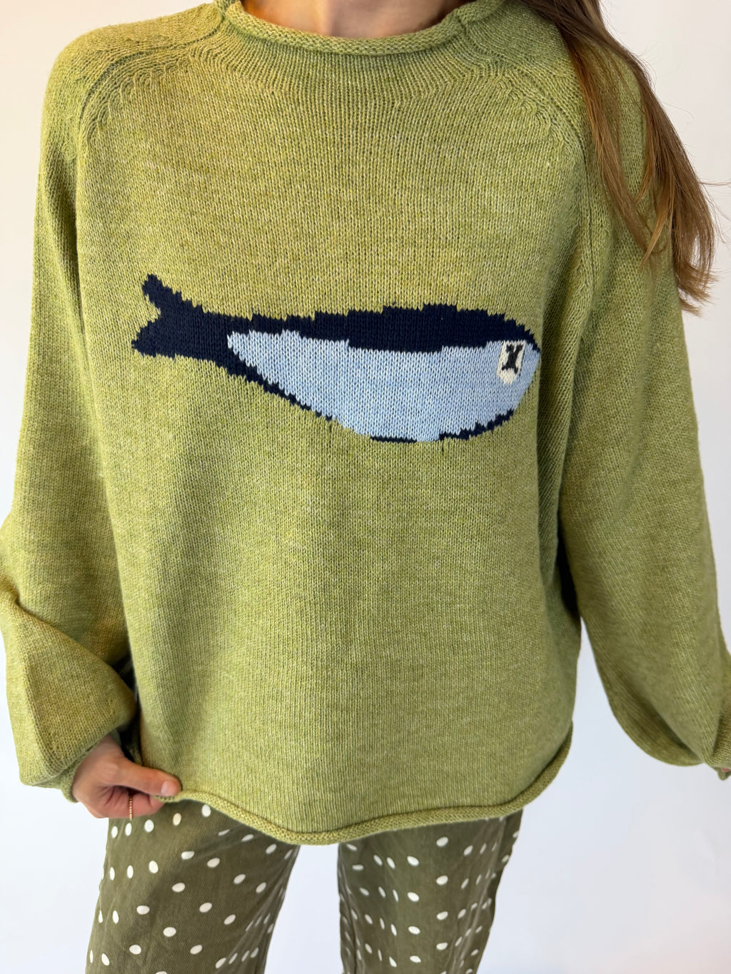 A person wearing a knitted sweater in a muted olive green color with a large fish motif across the chest in navy, light blue, and white. The sweater has a crew neck and long sleeves, paired with olive green pants with white polka dots.