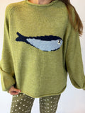 A person wearing a knitted sweater in a muted olive green color with a large fish motif across the chest in navy, light blue, and white. The sweater has a crew neck and long sleeves, paired with olive green pants with white polka dots.