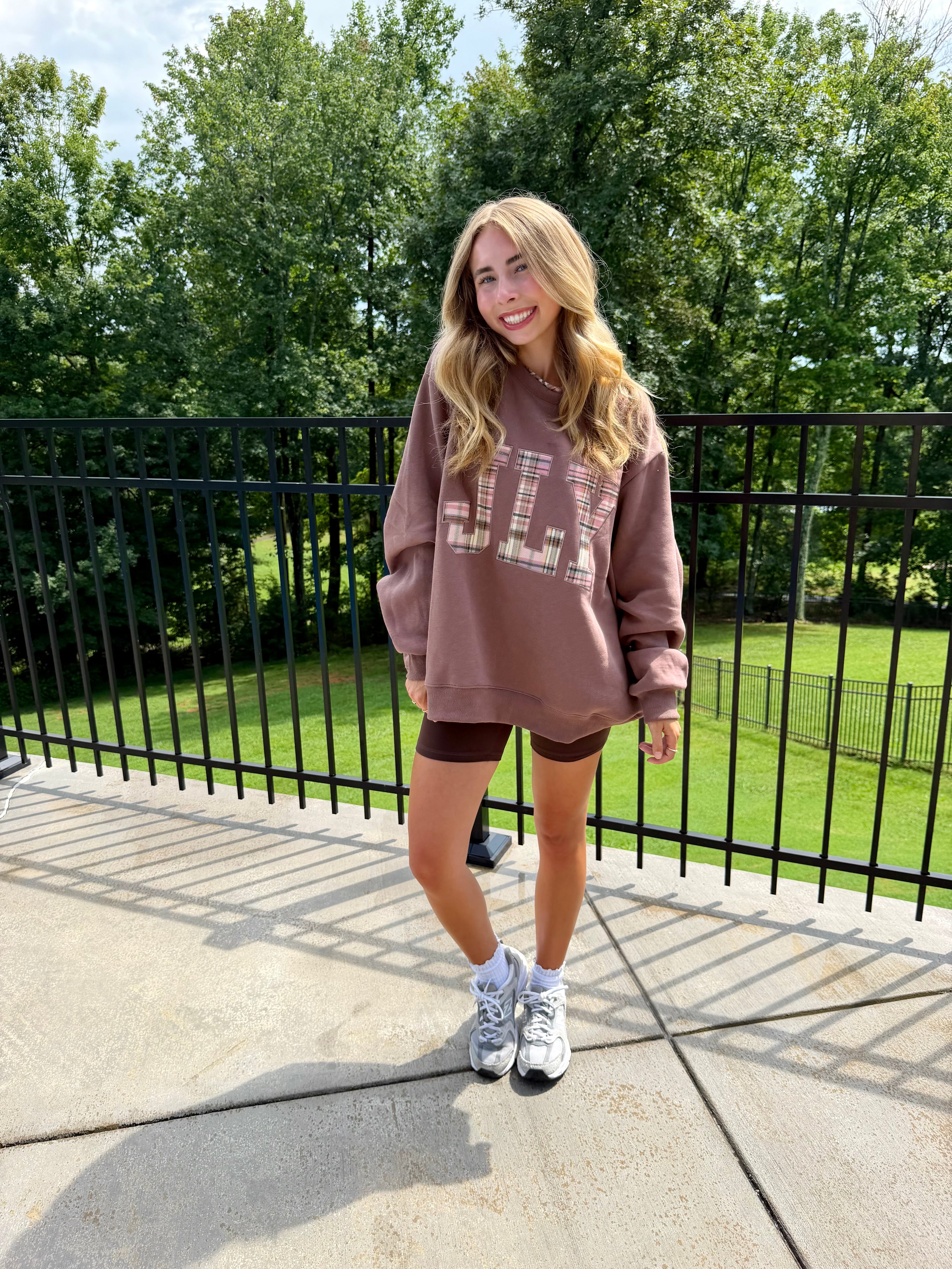 A woman stands on an outdoor concrete balcony wearing a dusty rose-brown oversized sweatshirt with a large graphic print on the front, paired with dark brown shorts and white sneakers. The background features a black metal railing and a lush green lawn with trees.