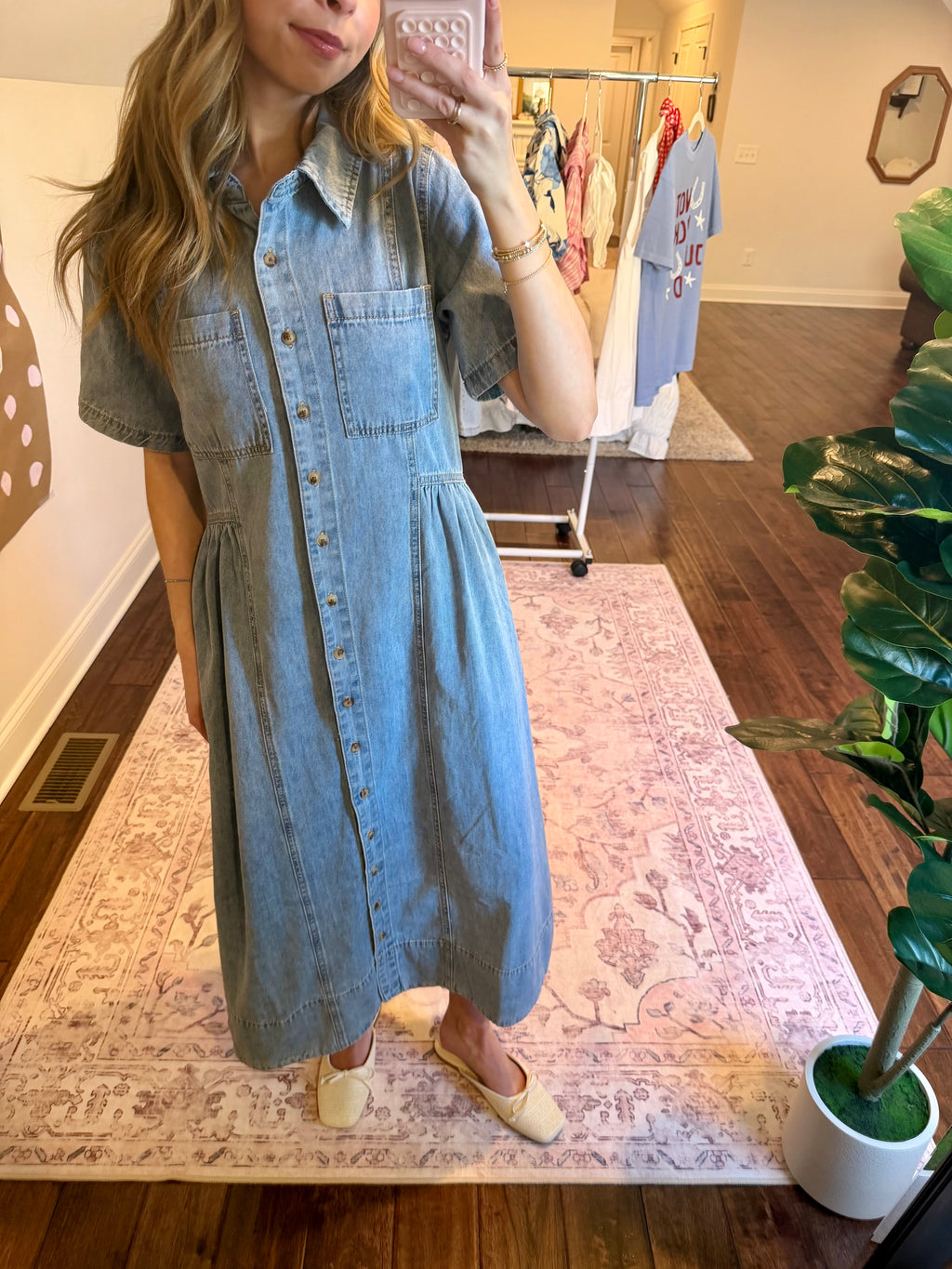 Person wearing a denim dress taking a mirror selfie in a room with a plant and clothing rack.