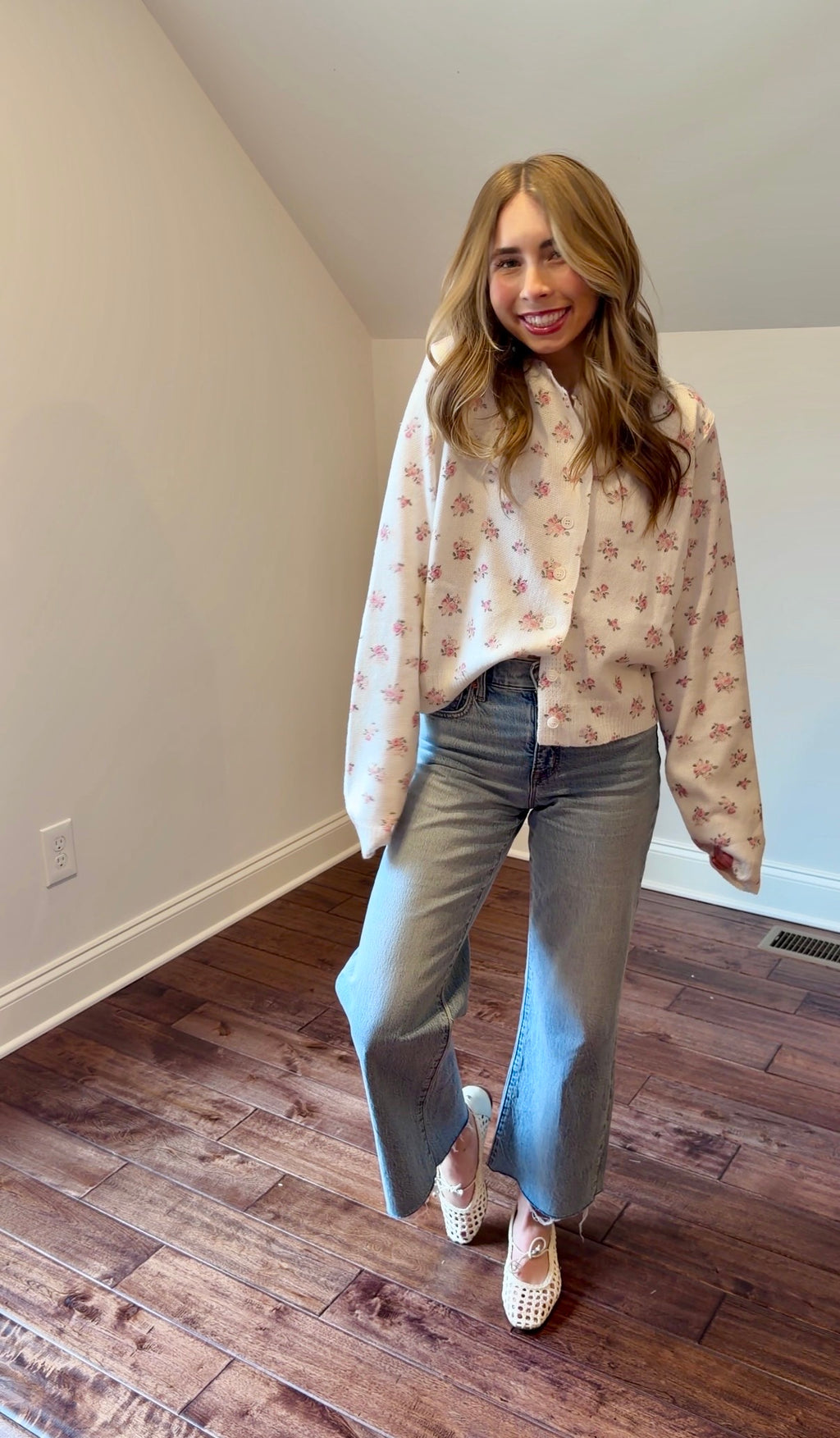 Woman wearing a floral cardigan and jeans in a room with wooden flooring.