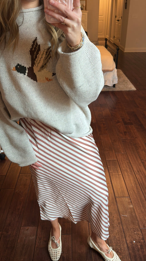 Person wearing a white sweater with a duck design and a brown and white striped skirt indoors.