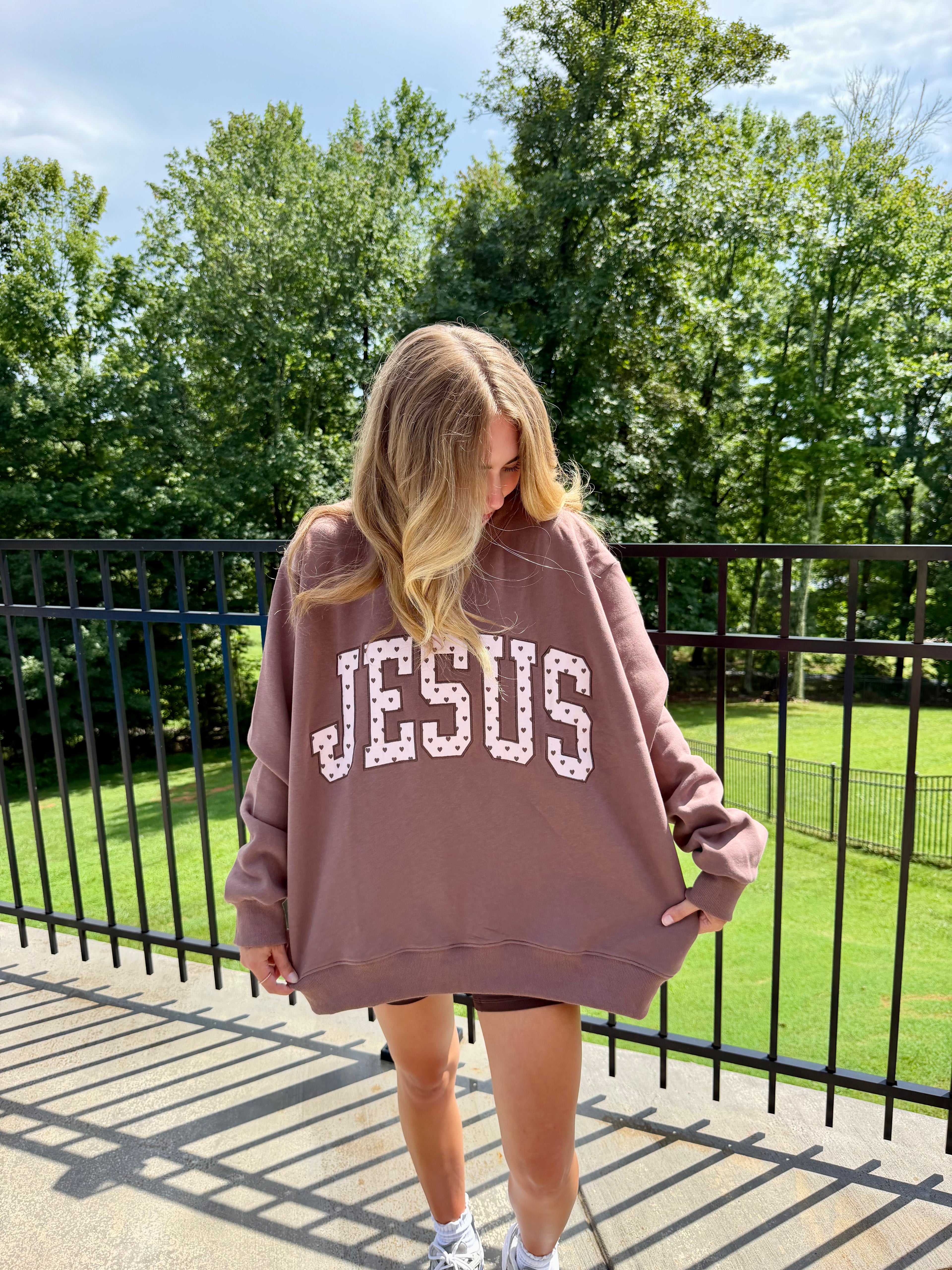 A woman wearing an oversized dusty rose sweatshirt with 'JESUS' printed across the front in white letters, standing outdoors on a balcony with trees and a fence behind her.