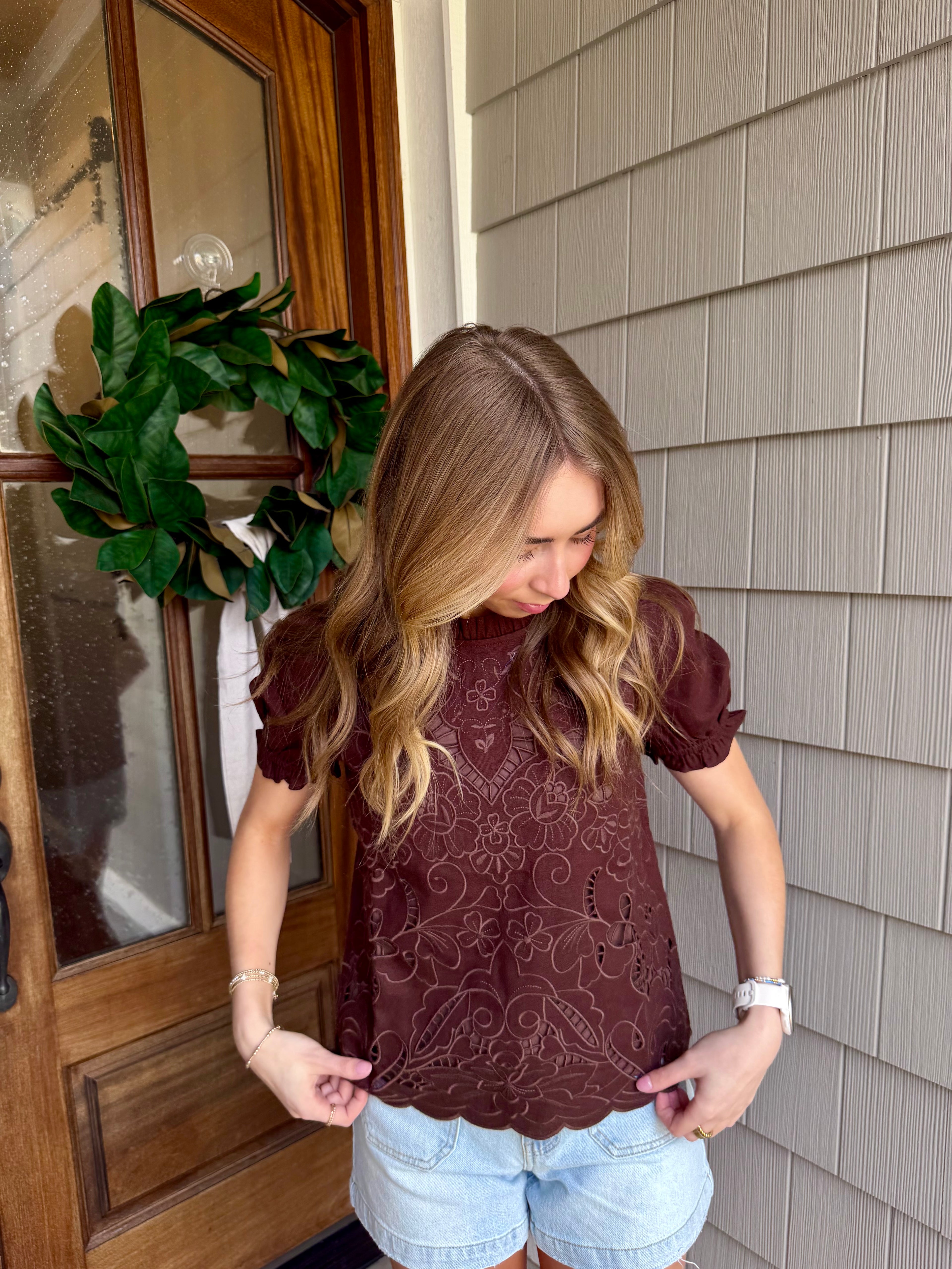 Woman wearing a brown patterned top and light blue shorts standing in front of a house door.