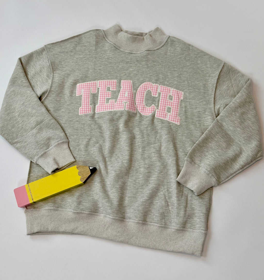 A light heather gray sweatshirt with the word 'TEACH' in pink gingham letters across the chest, styled flat on a light gray background with a yellow and pink pencil.